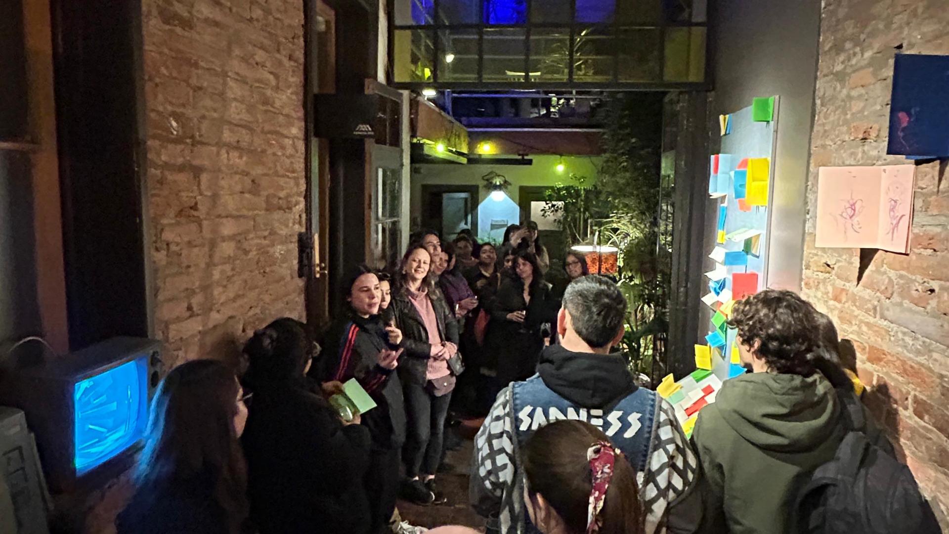 Grupo de personas reunidas en un pasillo interior con muros de ladrillo a la vista, observando una instalación compuesta por papeles de colores adheridos a la pared. La escena está iluminada con luces cálidas y al fondo se distingue un espacio con iluminación azul y verde.