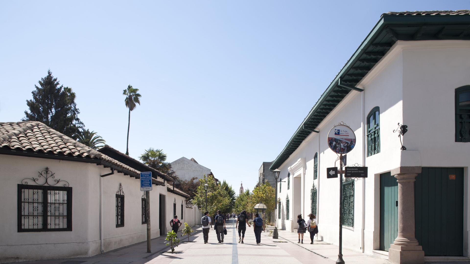 dos casas coloniales de color blanco con tejas de greda, ubicada una frente a la otra y separadas por un paseo petaonal donde transita gente.