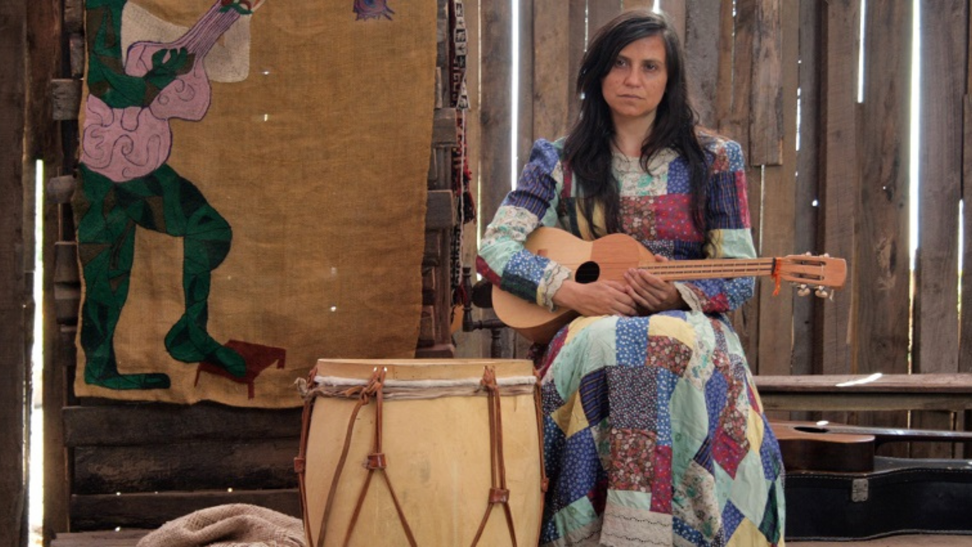 Francisca Gavilán personificando a Violeta Parra. Está sentada con un vestido coloreado y sosteniendo una guitarra y al lado un instrumento de percusión. En el fondo hay tablas de madera y lo que pareciera ser una obra (pintura o arpillera).
