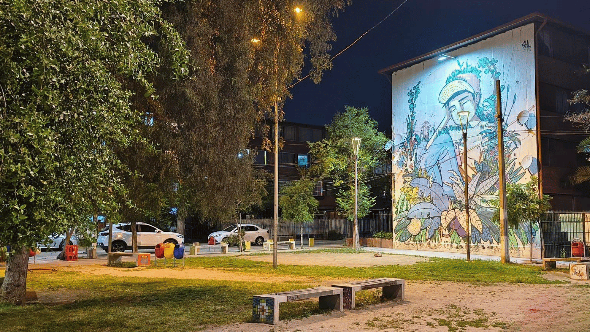 Imagen de plaza el progreso. Se observa un mural en el fondo, vegetación y mobiliarios propios de una plaza (arboles, pasto y bancas de cemento).