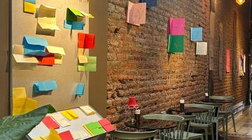 Pasillo interior con muro de ladrillo a la vista donde se exhiben hojas de papel de colores dobladas y colgadas. A la izquierda, un panel con papeles de tonos rojo, azul, amarillo y verde sobresaliendo. Hay mesas pequeñas con sillas verdes alineadas a lo largo del muro y una luz cálida al fondo del pasillo.